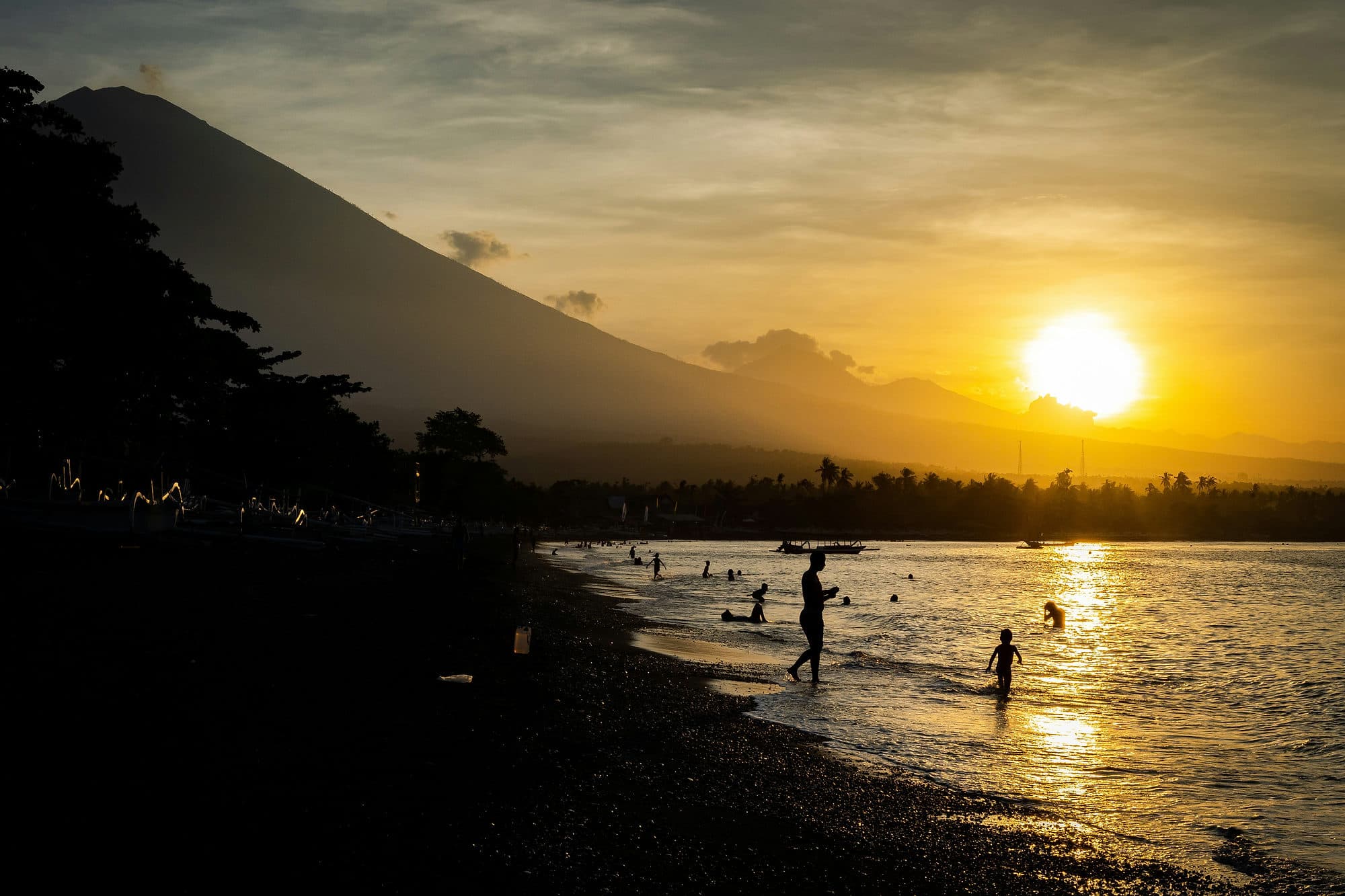 De zon zakt achter de vulkaan Agung terwijl mensen in zee zwemmen bij Amed