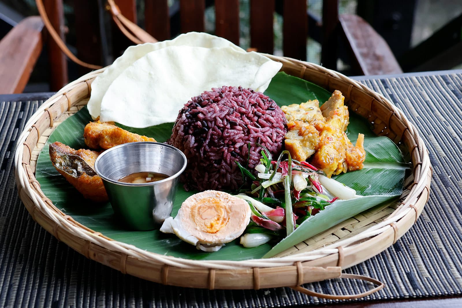 Een rieten schaal met zwarte rijst, kroepoek en sambal op een bananenblad