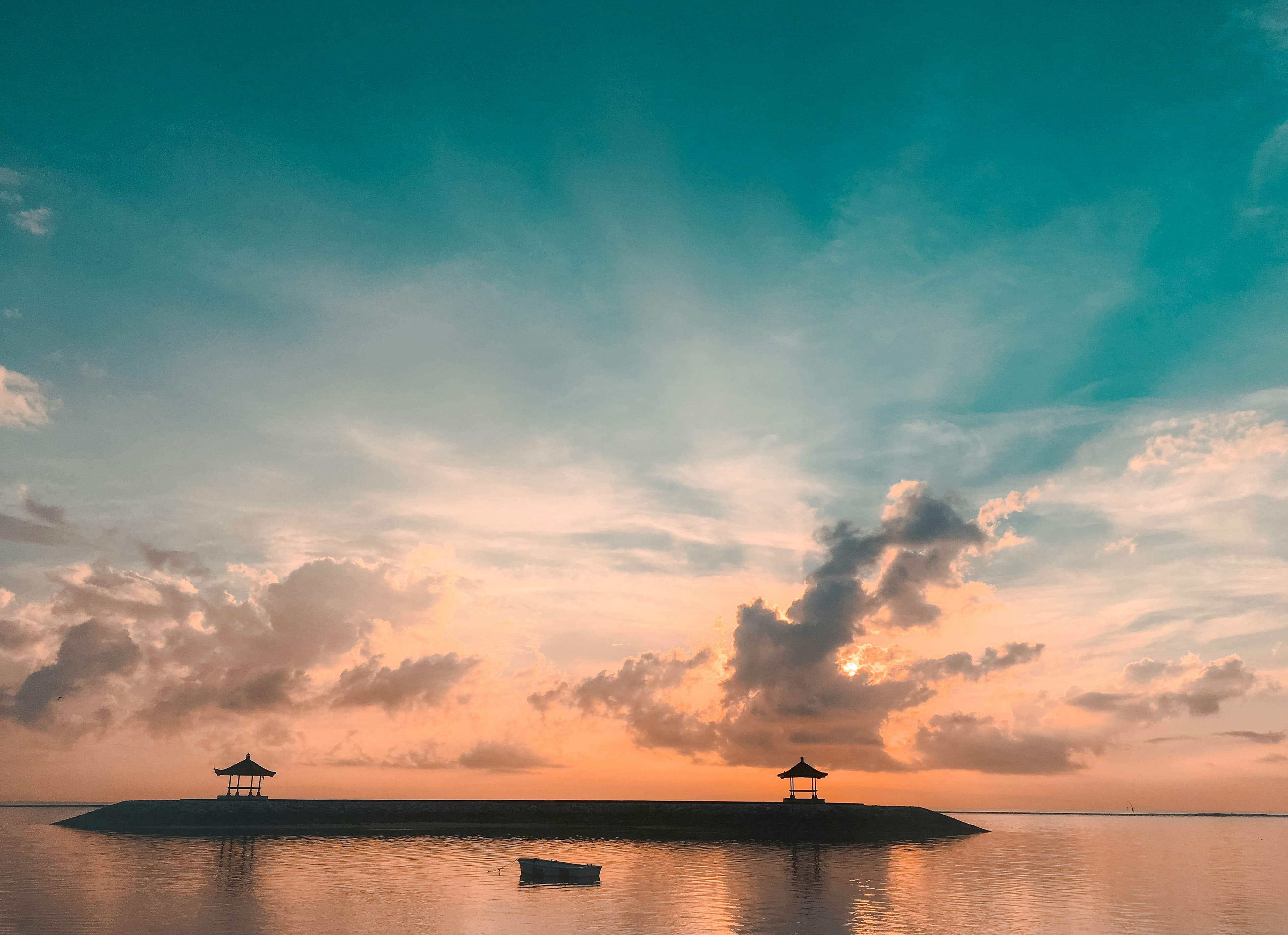 Twee Balinese paviljoentjes silhouet tegen een teal-oranje zonsopkomst boven kalme zee op Bali