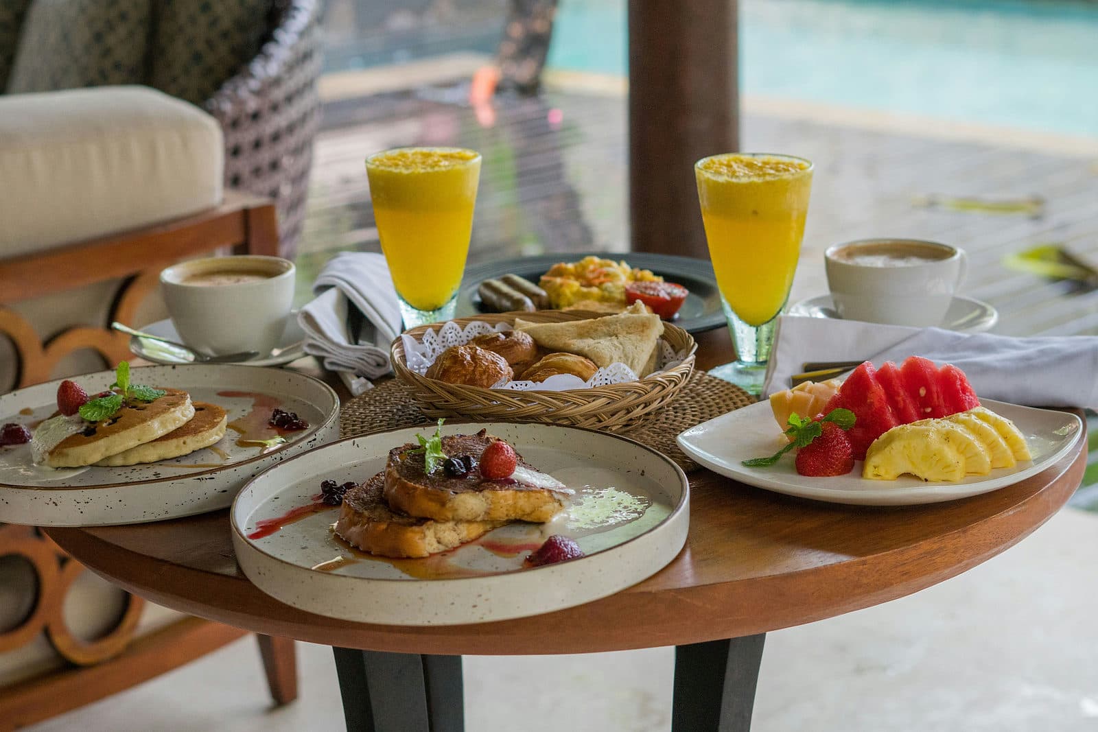 Ontbijt met pannenkoeken, fruit en verse sappen op een tafel bij een zwembad in Canggu