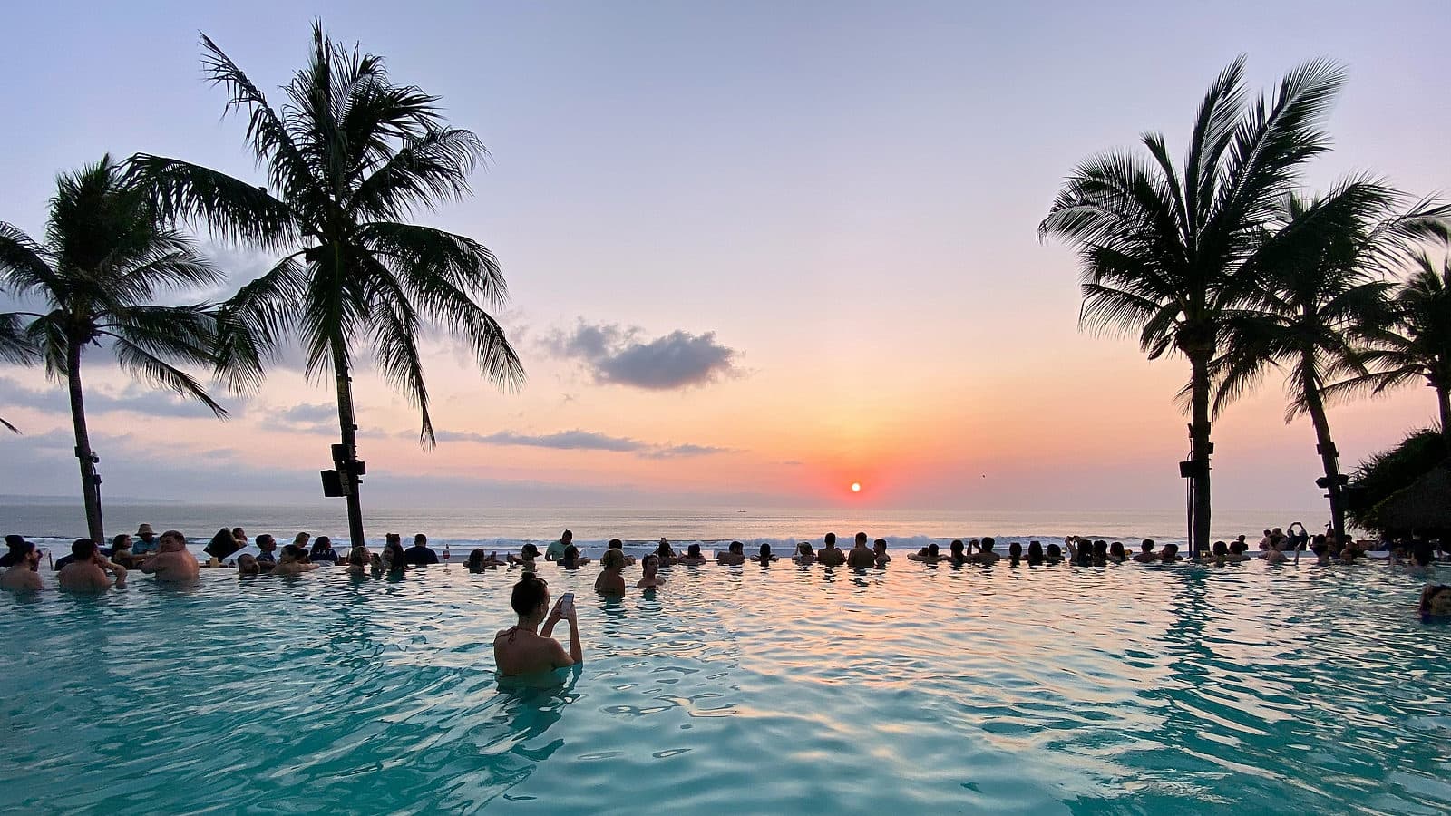 Mensen in een infinity pool aan het strand kijken naar de zonsondergang tussen de palmen