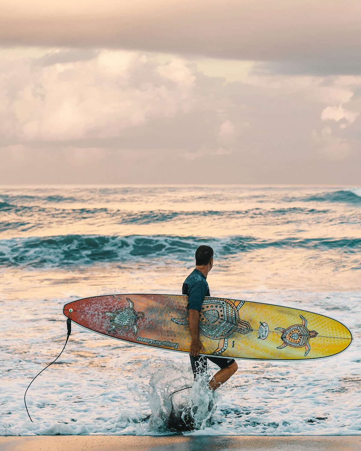 Een surfer loopt met een beschilderd longboard uit de branding het strand op