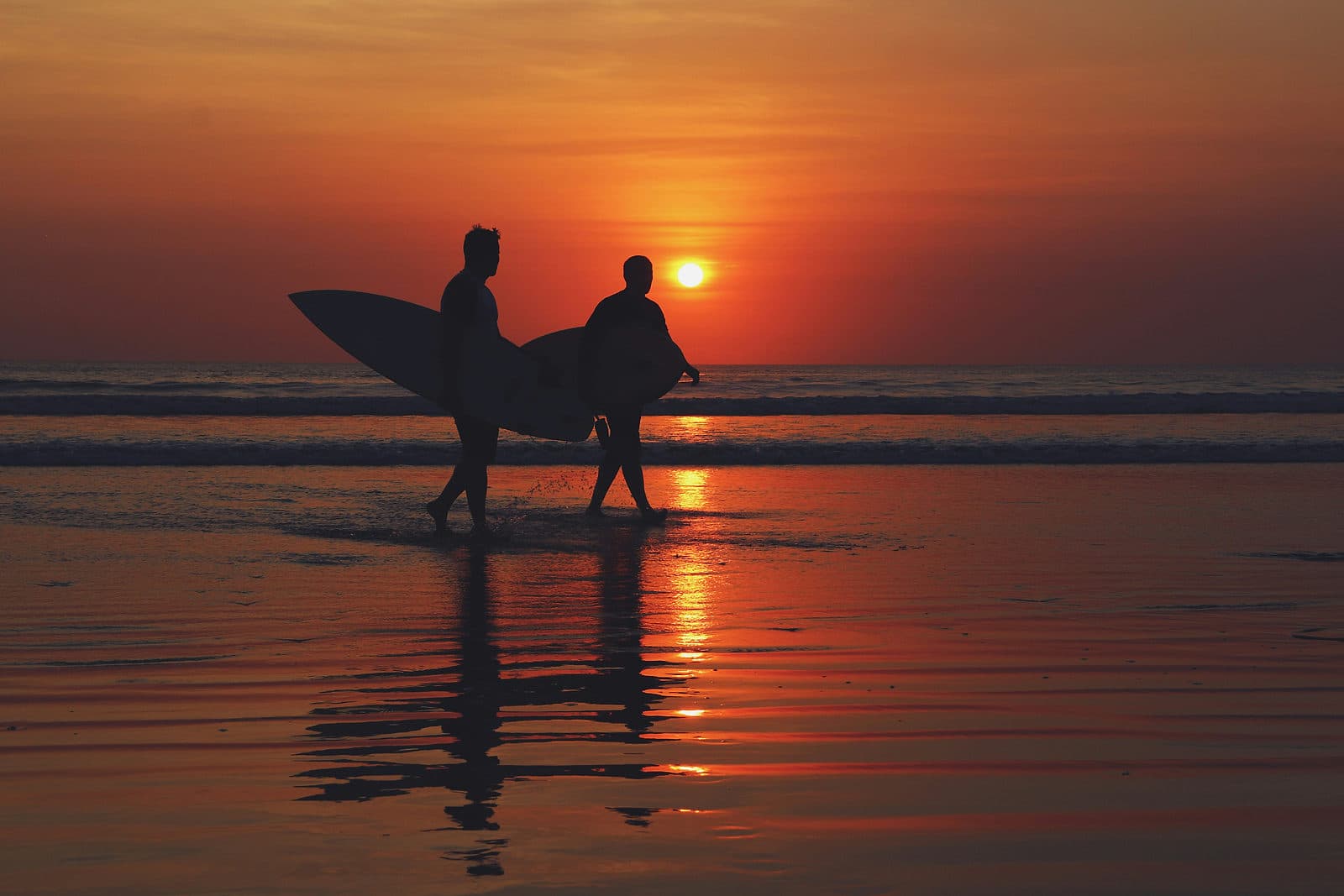 Twee surfers lopen in silhouet met hun board door het ondiepe water bij zonsondergang