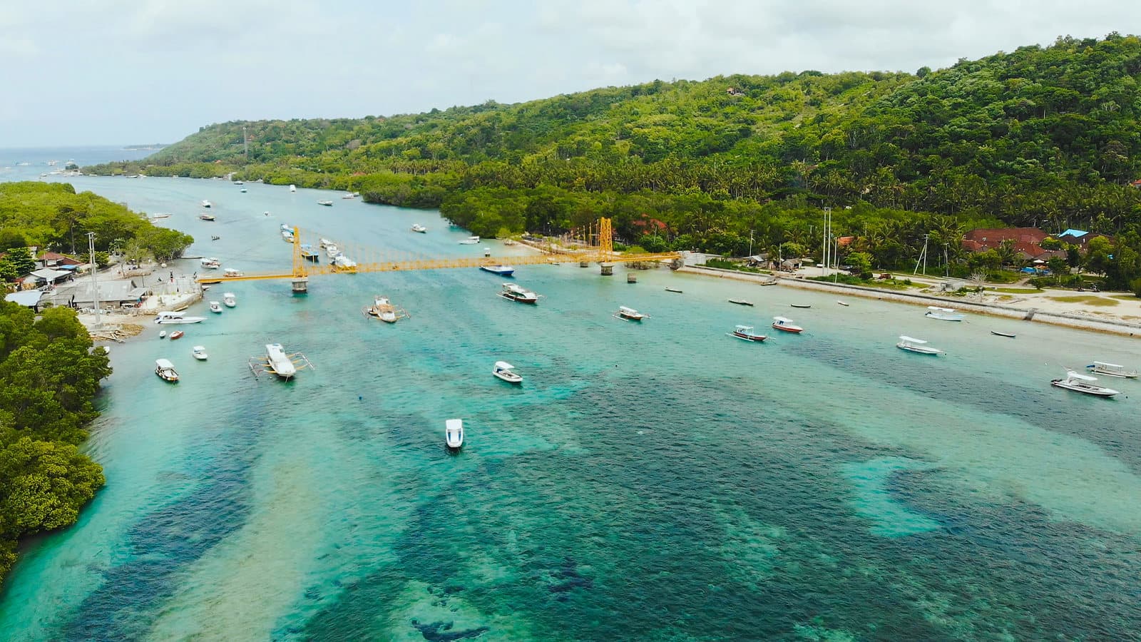 Luchtfoto van het kanaal tussen Nusa Lembongan en Nusa Ceningan, met de gele brug, ankerende bootjes en turkoois water boven het rif