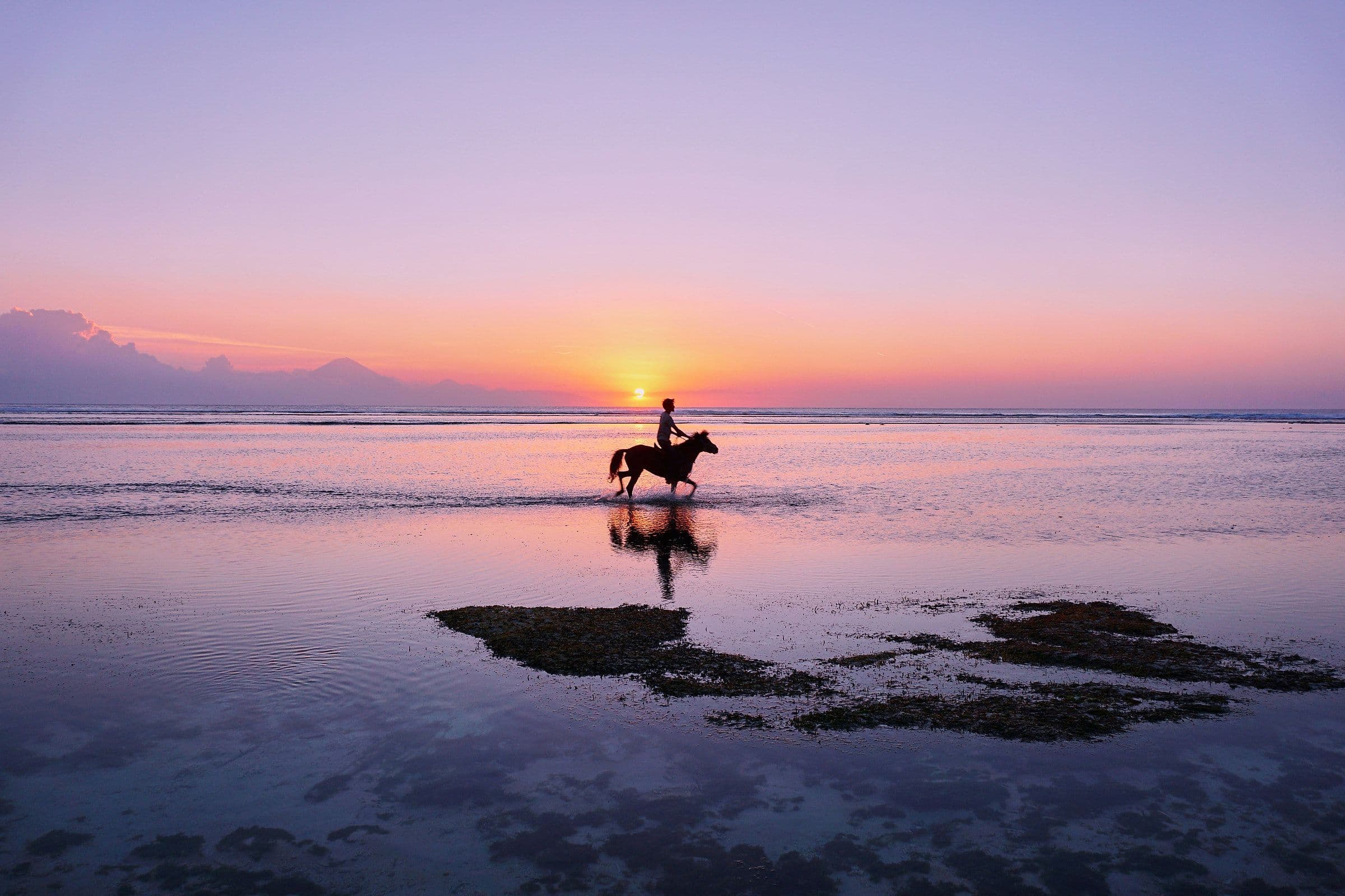 Iemand rijdt te paard door het ondiepe water voor de kust van een Gili-eiland terwijl de zon ondergaat