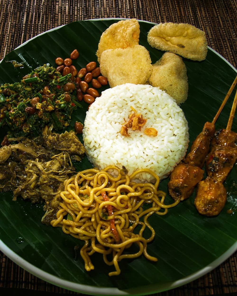 Een bord nasi campur met saté en groenten op een bananenblad