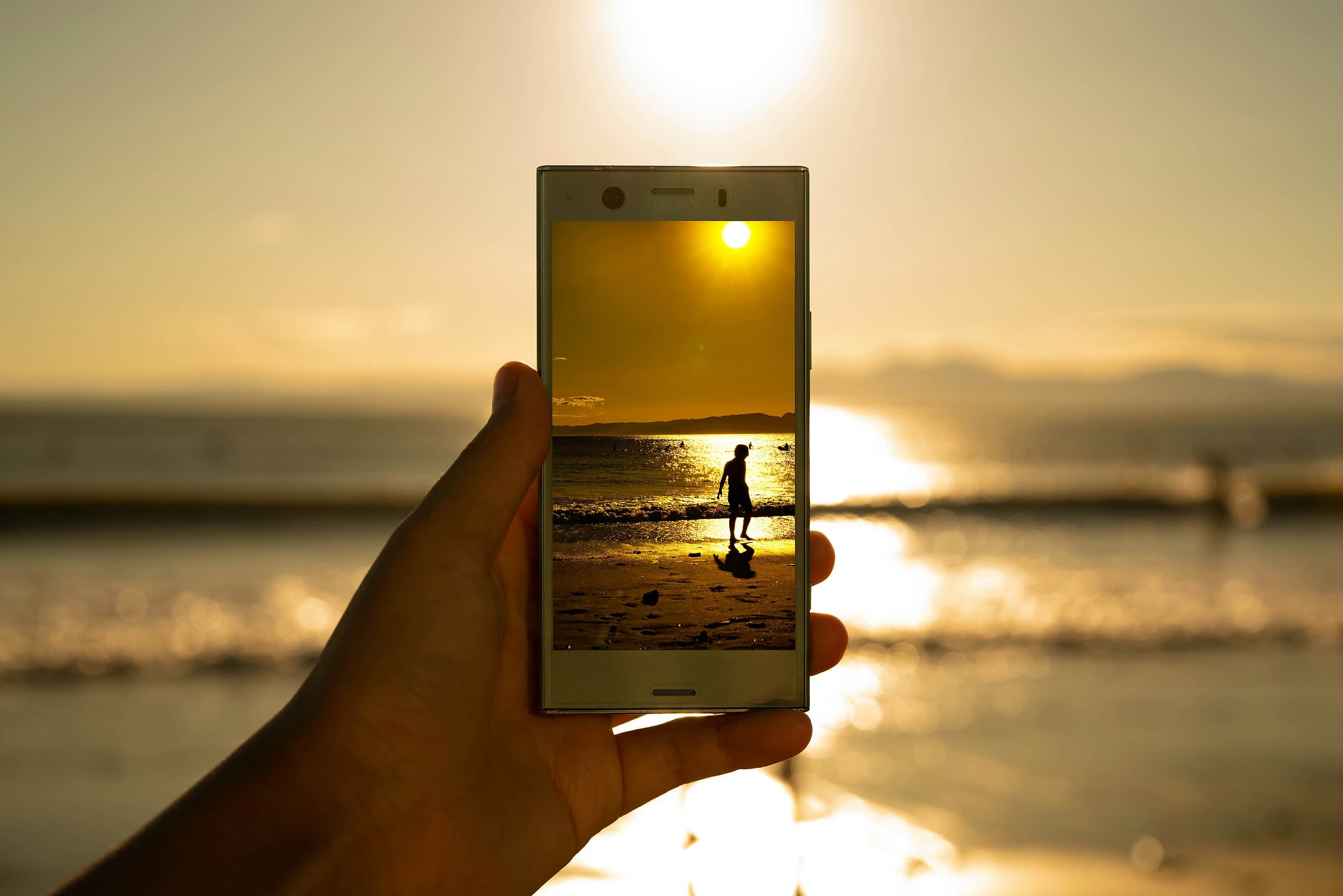 Een hand houdt een telefoon omhoog bij het strand in de avondzon, met op het scherm de wandelaar aan de waterlijn
