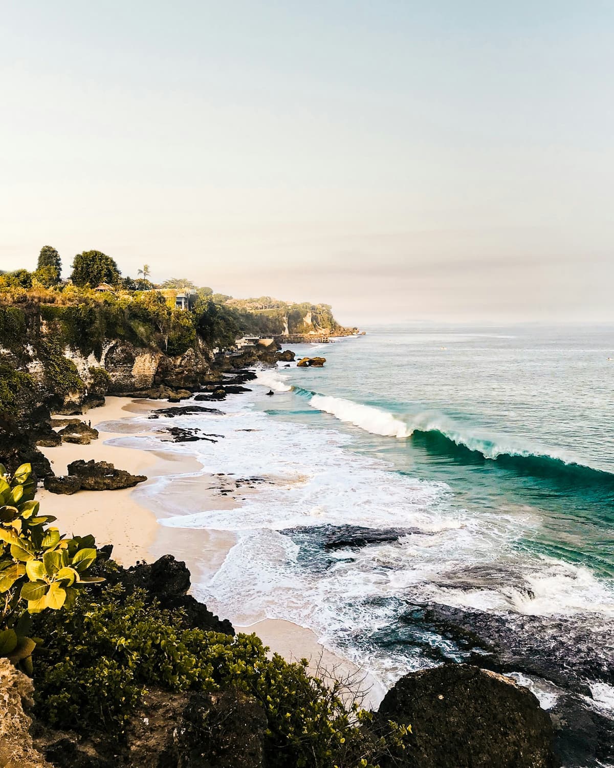 Lang wit strand met brekende golven aan de voet van een begroeide kalksteenklif op Bali