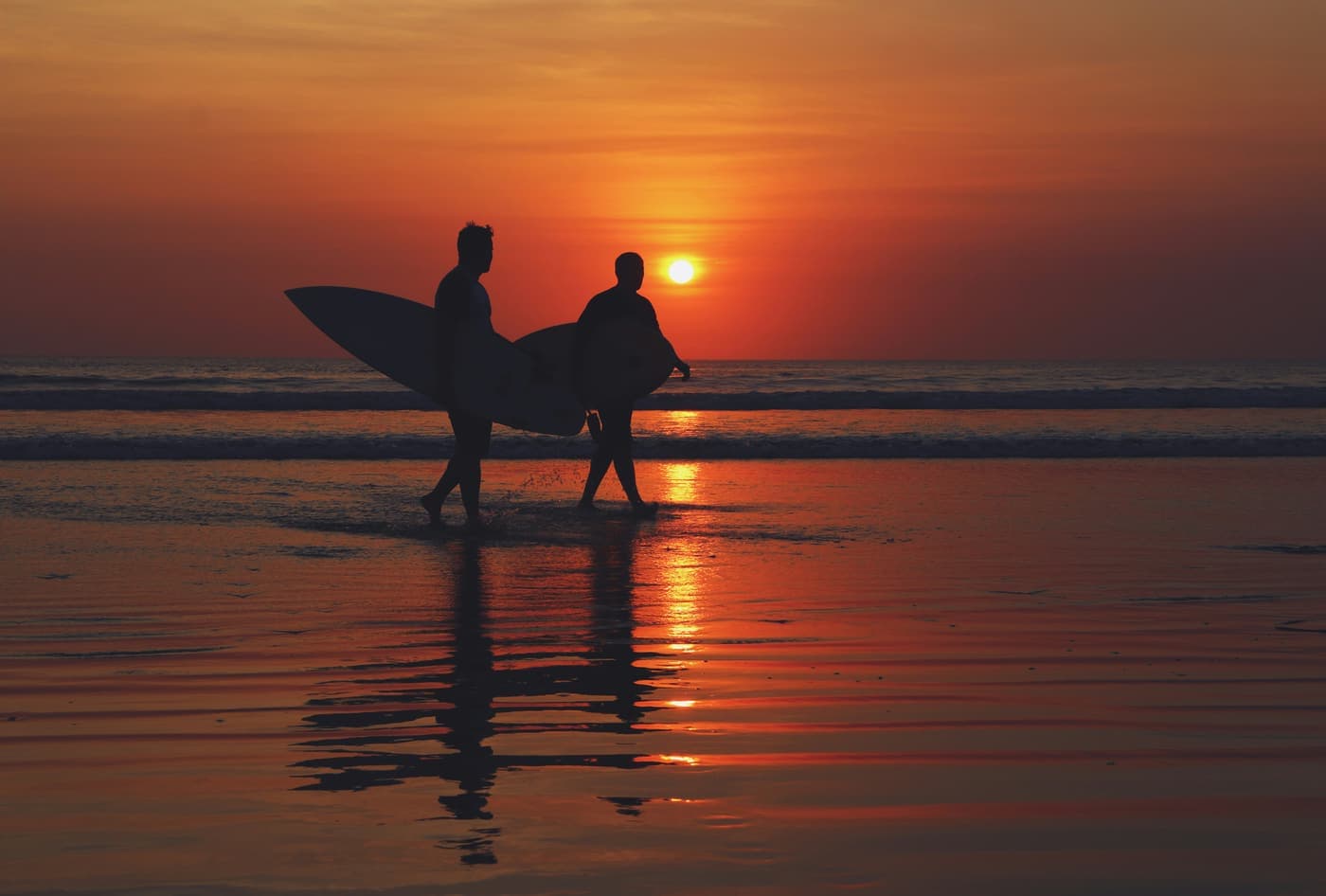 Twee surfers lopen in silhouet over het natte zand terwijl de zon in zee zakt