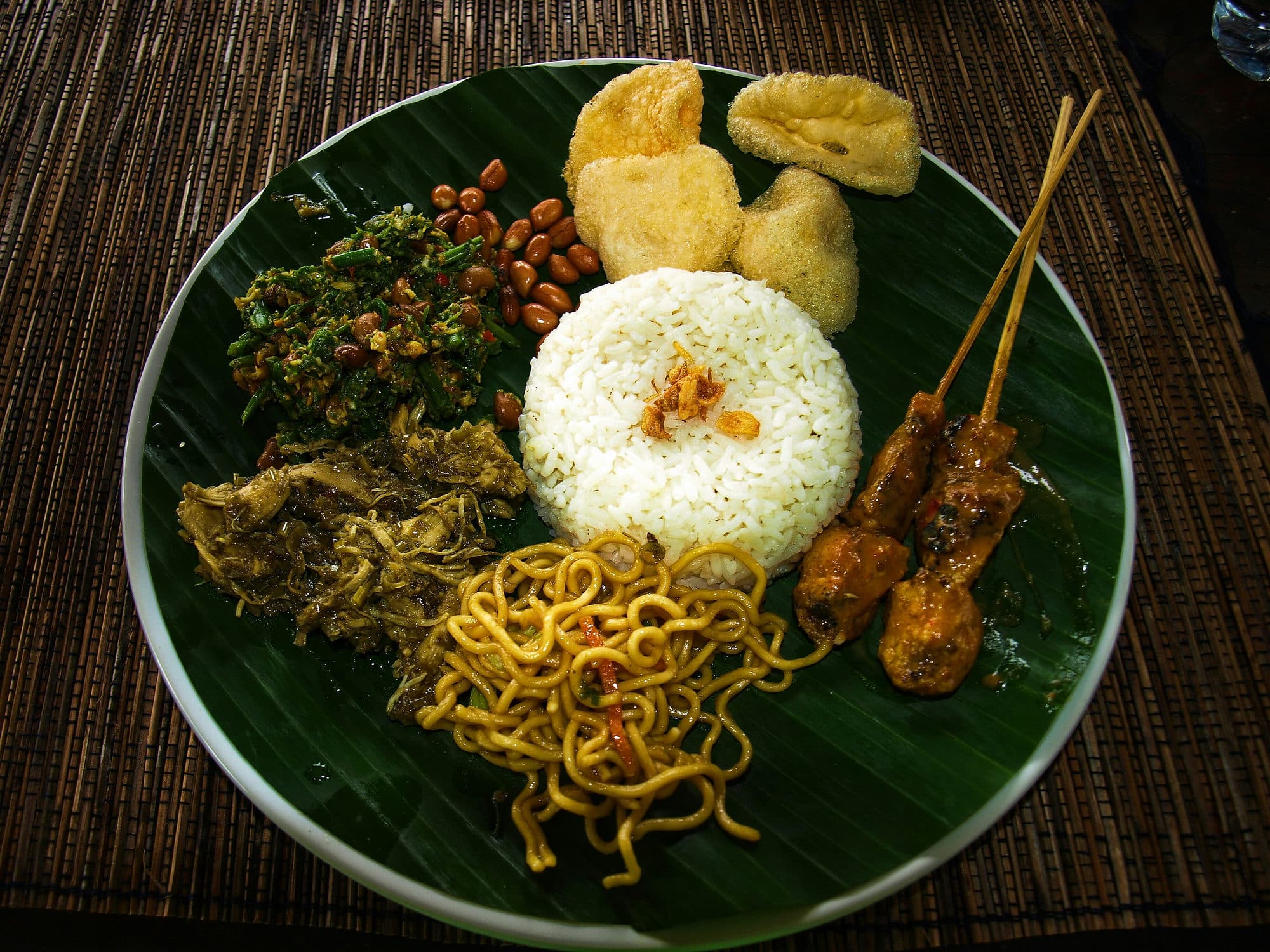 Een bord nasi campur op een bananenblad, met rijst, sate, sambal en kroepoek