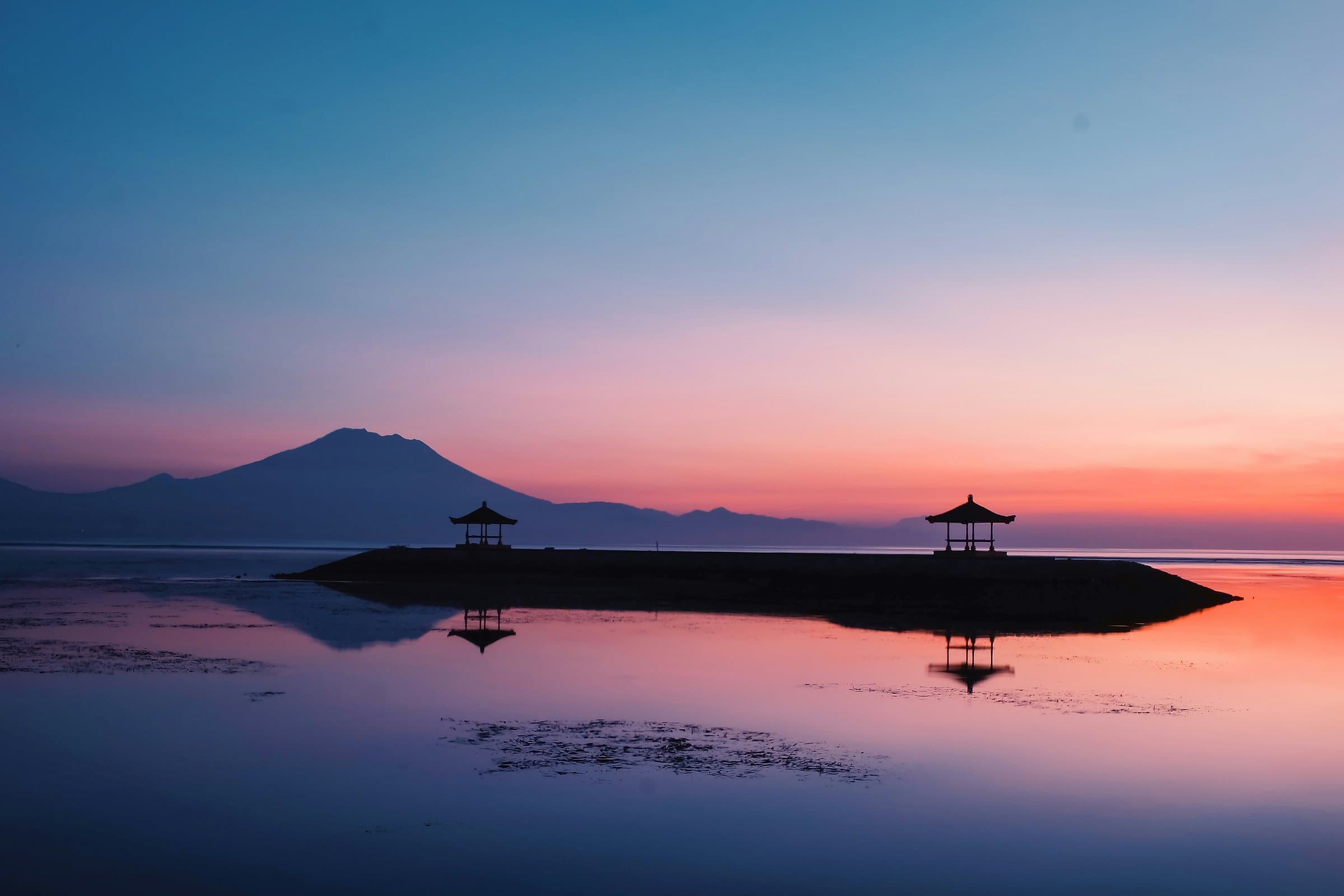 Twee bales op een stenen golfbreker van Sanur in het eerste licht, met een vulkaan aan de horizon boven spiegelglad water