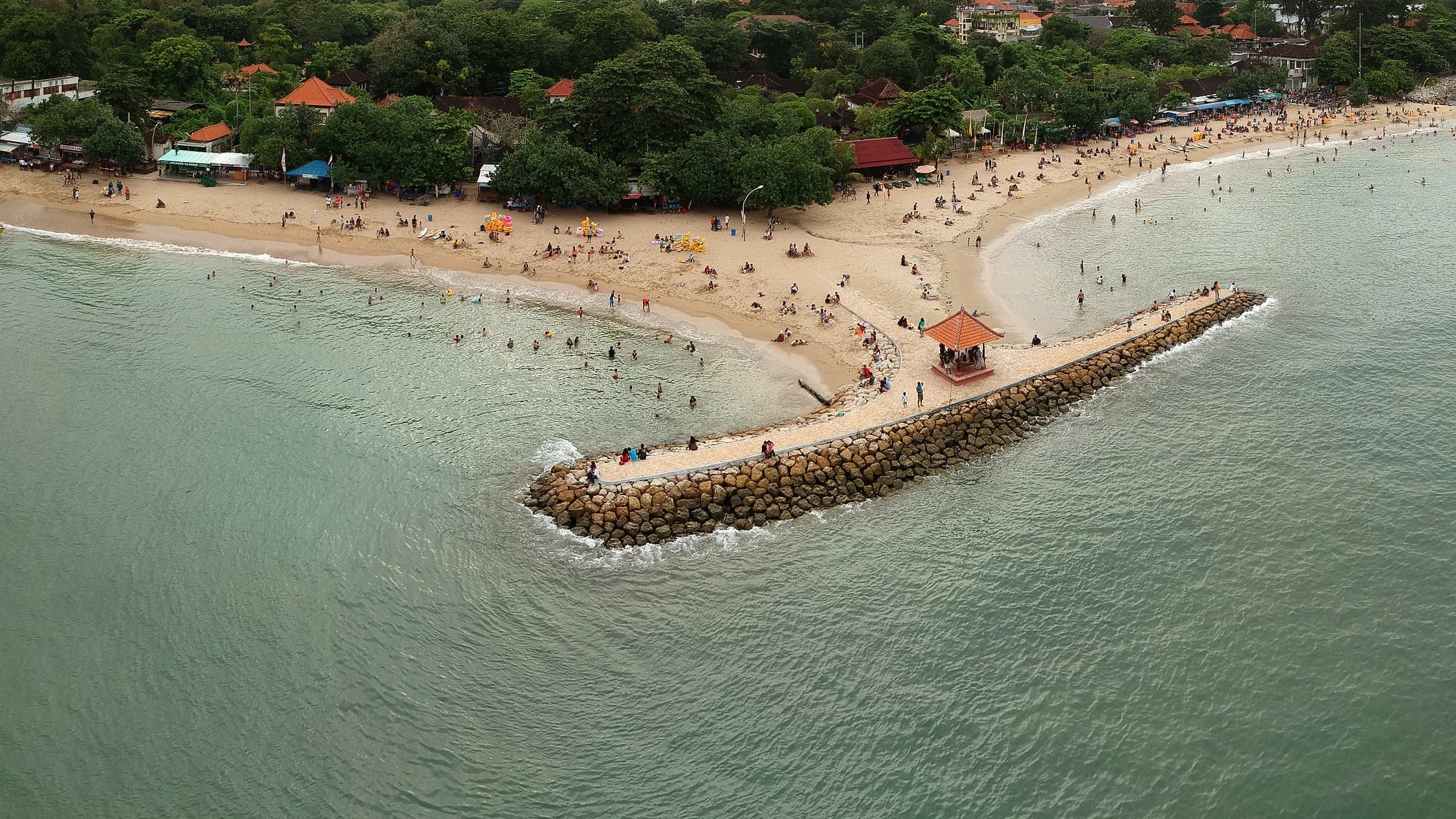 Luchtfoto van het strand van Sanur met een stenen golfbreker, een bale op het uiteinde en zwemmers in de kalme zee