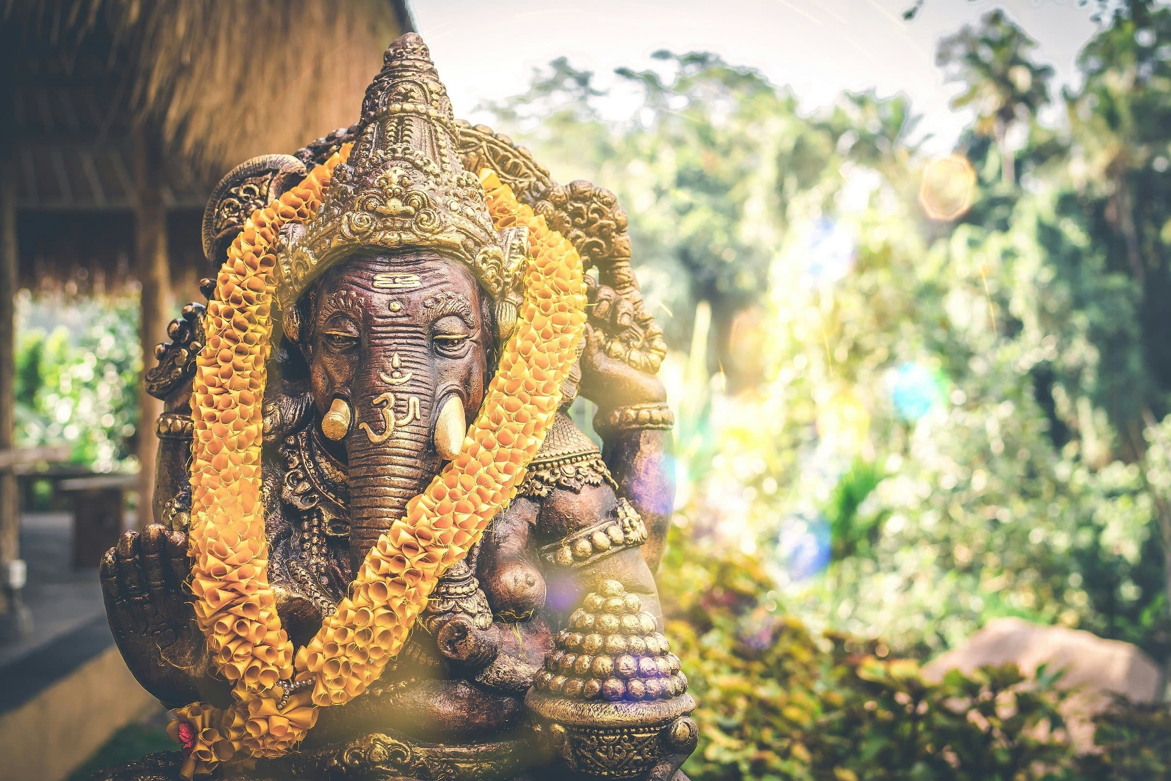 Stenen Ganeshabeeld met een gele bloemenslinger om, in het groen bij een Balinese tempel